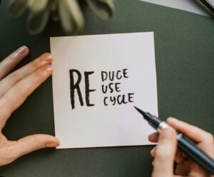 Close-up of hands writing a motivational 'Reduce Reuse Recycle' note on paper with a pen, surrounded by office items.